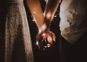 A warm and intimate close-up of a couple holding hands adorned with glowing string lights.